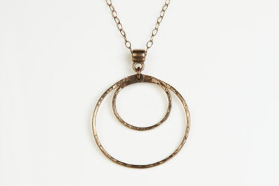 Double Happiness Bronze Circle Pendant - shown hanging on a white background. The two handmade, hammered, concentric circles on a rose gold chain reflect with lovely contrasting warm bronze tones. The two nested circles rustic look is complimented by the hypoallergenic gold fill chain and handmade wire wrapped clasp. Together, the Double Happiness Bronze Circle Pendant is a simple necklace to wear everyday, or on the best of occasions. Bronze 8th anniversary gifts for her