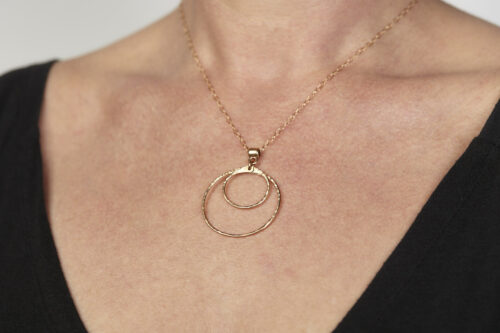 Double Happiness Bronze Circle Pendant Necklace - shown worn on a woman wearing a black scoop neck sweater. The two handmade, hammered, concentric circles on a rose gold chain reflect with lovely contrasting warm bronze tones. The two nested circles rustic look is complimented by the hypoallergenic gold fill chain and handmade wire wrapped clasp. Together, the Double Happiness Bronze Circles Pendant Necklace is a simple necklace to wear everyday, or on the best of occasions. Bronze 8th anniversary gifts for her