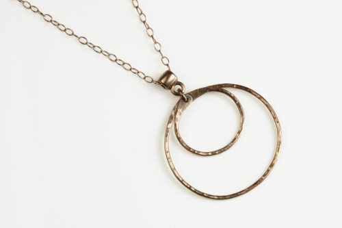 Double Happiness Bronze Circle Pendant Necklace - shown laying on a white background. The two handmade, hammered, concentric circles on a rose gold chain reflect with lovely contrasting warm bronze tones. The two nested circles rustic look is complimented by the hypoallergenic gold fill chain and handmade wire wrapped clasp. Together, the Double Happiness Bronze Circles Pendant Necklace is a simple necklace to wear everyday, or on the best of occasions. Bronze 8th anniversary gifts for her