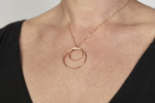 Double Happiness Copper Circle Pendant - two handmade, hammered, concentric circles on a rose gold chain reflecting with a lovely contrasting warm coppery tone. Worn by a woman with a black scoop neck sweater. Copper gifts for wife