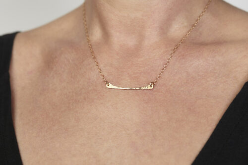 Simple Chic - Bronze Horizontal Bar Necklace displayed worn on a woman wearing a black scoop neck sweater. The uniquely minimalist Bronze curved bar necklace displays its hand forged textures in the warm light. The gold filled chain matches the warm tones of the Bronze showcasing its simple elegance. 8th anniversary gifts