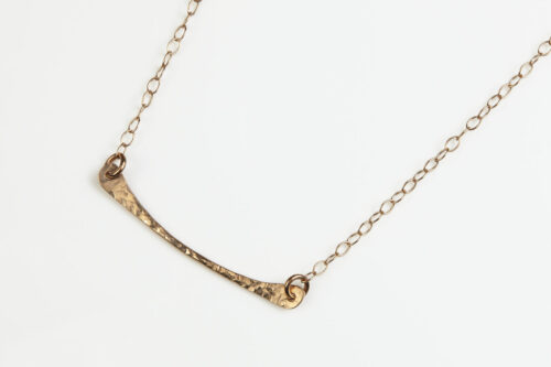 Simple Chic - Bronze Bar Pendant displayed laying on a white background. The uniquely minimalist Bronze curved bar necklace displays its hand forged textures in the warm light. The gold filled chain matches the warm tones of the Bronze showcasing its simple elegance. 8th anniversary gifts