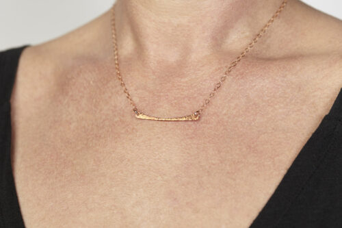 Simple Chic - Horizontal Bar Necklace displayed worn by a woman with a black v-neck sweater. The uniquely minimalist copper horizontal bar necklace displays its hand forged textures in the warm light. Rose gold filled chain matches the warm tones of the copper showcasing its simple elegance. Copper anniversary gifts for her
