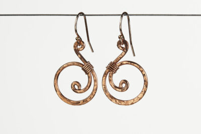 Tiny Swirls - Copper Swirl Earrings displayed hanging on a white background. These copper drop hoop earrings have a wire wrapped copper detail that creates the closed circle of the hoops with a tiny spiral inside the hoop to provide the cute focal point of the earrings. Delicately hammered facets reflect the light of the copper. The rose gold fill ear wires match the warm tones of the copper perfectly. Small and light enough for every day, but sophisticated enough for a fancy night out, the Tiny Swirls copper swirl earrings are perfect for every occasion. Copper anniversary gifts for her