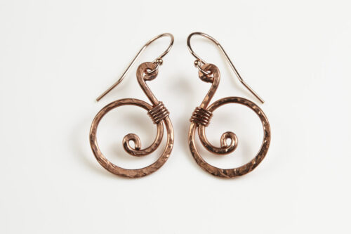 Tiny Swirls - Copper Spiral Earrings displayed laying on a white background. These copper drop hoop earrings have a wire wrapped copper detail that creates the closed circle of the hoops with a tiny spiral inside the hoop to provide the cute focal point of the earrings. Delicately hammered facets reflect the light of the copper. The rose gold fill ear wires match the warm tones of the copper perfectly. Small and light enough for every day, but sophisticated enough for a fancy night out, the Tiny Swirls copper spiral earrings are perfect for every occasion. Copper gifts for her