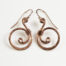 Tiny Swirls - Copper Spiral Earrings displayed laying on a white background. These copper drop hoop earrings have a wire wrapped copper detail that creates the closed circle of the hoops with a tiny spiral inside the hoop to provide the cute focal point of the earrings. Delicately hammered facets reflect the light of the copper. The rose gold fill ear wires match the warm tones of the copper perfectly. Small and light enough for every day, but sophisticated enough for a fancy night out, the Tiny Swirls copper spiral earrings are perfect for every occasion. Copper gifts for her