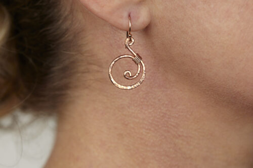 Tiny Swirls - Copper Spiral Earrings displayed laying on a white background. These copper drop hoop earrings have a wire wrapped copper detail that creates the closed circle of the hoops with a tiny spiral inside the hoop to provide the cute focal point of the earrings. Delicately hammered facets reflect the light of the copper. The rose gold fill ear wires match the warm tones of the copper perfectly. Small and light enough for every day, but sophisticated enough for a fancy night out, the Tiny Swirls copper spiral earrings are perfect for every occasion. Copper gifts for her