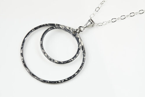 Double Happiness Aluminum Circles Necklace - shown laying on a white background. The two handmade, hammered, concentric circles on a sterling silver chain reflect with lovely contrasting aluminum tones. The two nested circles rustic look is complimented by the hypoallergenic sterling silver chain and handmade wire wrapped clasp. Together, the Double Happiness Aluminum Circles Necklace is a simple necklace to wear everyday, or perfect for a night on the town.