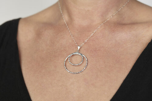Double Happiness Aluminum Circles Necklace - shown laying on a white background. The two handmade, hammered, concentric circles on a sterling silver chain reflect with lovely contrasting aluminum tones. The two nested circles rustic look is complimented by the hypoallergenic sterling silver chain and handmade wire wrapped clasp. Together, the Double Happiness Aluminum Circles Necklace is a simple necklace to wear everyday, or perfect for a night on the town.