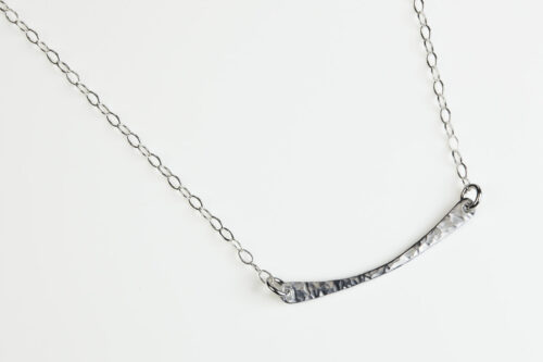 Simple Chic - Aluminum Bar Necklace displayed laying on a white background. The uniquely minimalist Aluminum curved bar necklace displays its hand forged textures in the warm light. The sterling silver chain matches the cool tones of the Aluminum showcasing its simple elegance.
