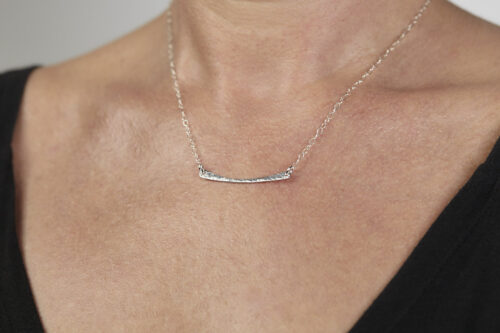 Simple Chic - Aluminum Horizontal Bar Necklace displayed laying on a white background. The uniquely minimalist Aluminum curved bar necklace displays its hand forged textures in the warm light. The sterling silver chain matches the cool tones of the Aluminum showcasing its simple elegance.