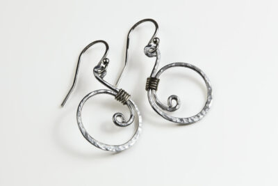 Tiny Swirls - Aluminum Spiral Earrings displayed laying on a white background. These Aluminum drop hoop earrings have a wire wrapped Aluminum detail that creates the closed circle of the hoops with a tiny spiral inside the hoop to provide the cute focal point of the earrings. Delicately hammered facets reflect the light of the Aluminum. The sterling silver ear wires match the warm tones of the Aluminum perfectly. Small and light enough for every day, but sophisticated enough for a fancy night out, the Tiny Swirls - Aluminum Spiral Earrings are perfect for every occasion.