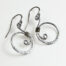 Tiny Swirls - Aluminum Spiral Earrings displayed laying on a white background. These Aluminum drop hoop earrings have a wire wrapped Aluminum detail that creates the closed circle of the hoops with a tiny spiral inside the hoop to provide the cute focal point of the earrings. Delicately hammered facets reflect the light of the Aluminum. The sterling silver ear wires match the warm tones of the Aluminum perfectly. Small and light enough for every day, but sophisticated enough for a fancy night out, the Tiny Swirls - Aluminum Spiral Earrings are perfect for every occasion.